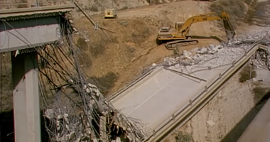 A partially collapsed highway bridge lies in ruins, with twisted rebar and broken concrete visible. Construction equipment, including excavators and bulldozers, are positioned nearby on a dirt road as part of a demolition or reconstruction effort. The scene is set in a hilly, dry landscape.